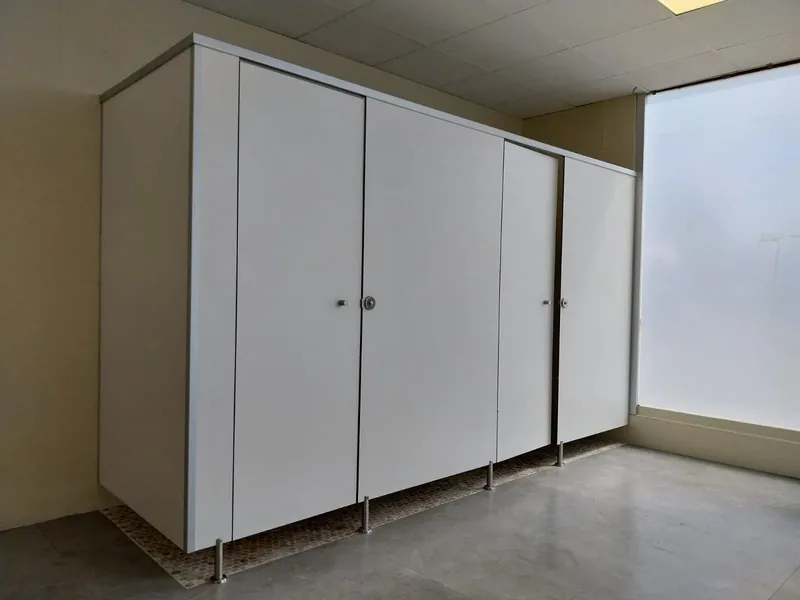 Cabinas sanitarias blancas con perfil de coronación de aluminio y pies regulables, junto a una ventana