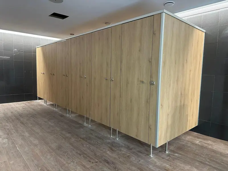 Cabinas sanitarias fenólicas en acabado roble, con perfil de coronación y sobre pies
