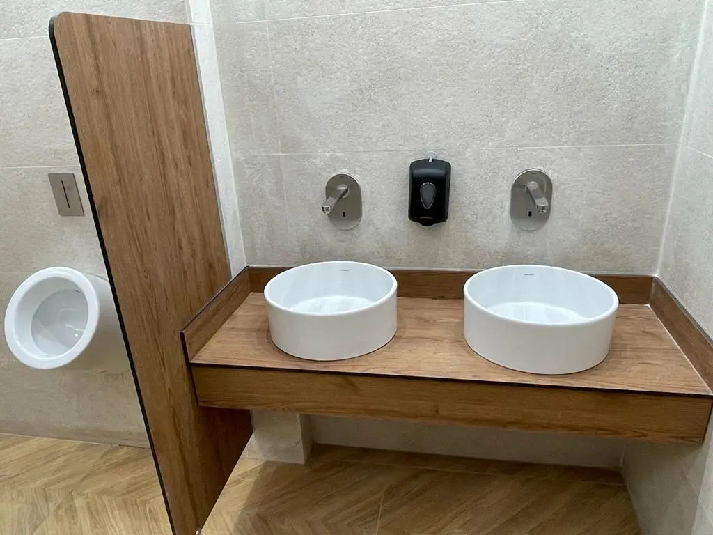 Dos lavabos de vaso blancos y redondos instalados sobre una encimera de madera en una configuración de montaje de vaso, con grifería de pared y un separador de privacidad en un baño con iluminación natural - Bellum Logistics