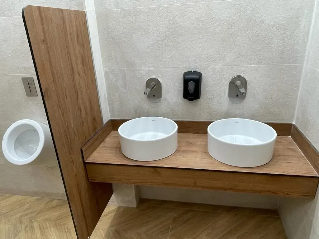 Separador de compacto fenólico en acabado madera entre un urinario y una encimera con dos lavabos, a juego