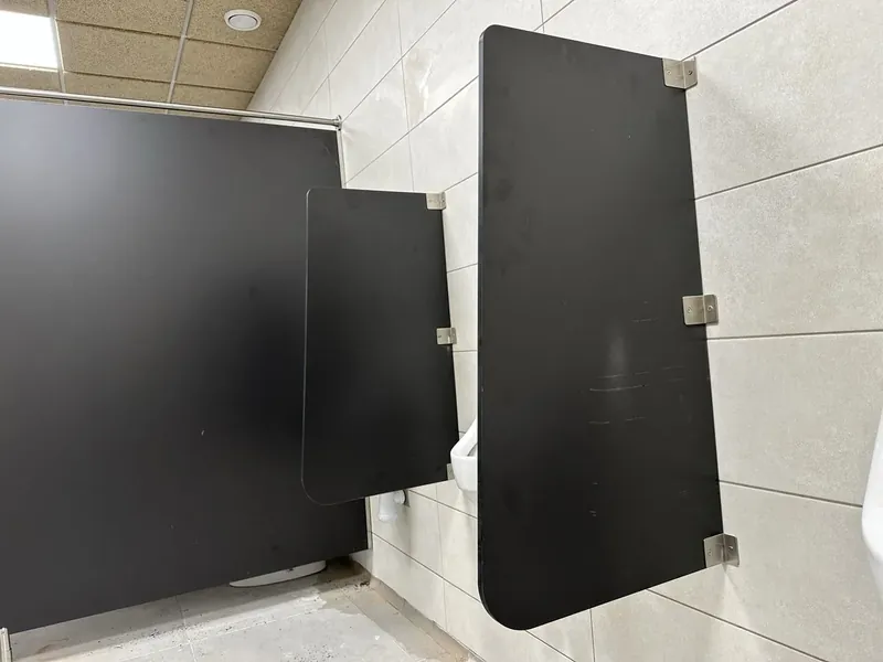 Separador de urinario negro sujeto a la pared con pinzas, junto a cabinas sanitarias del mismo compacto negro