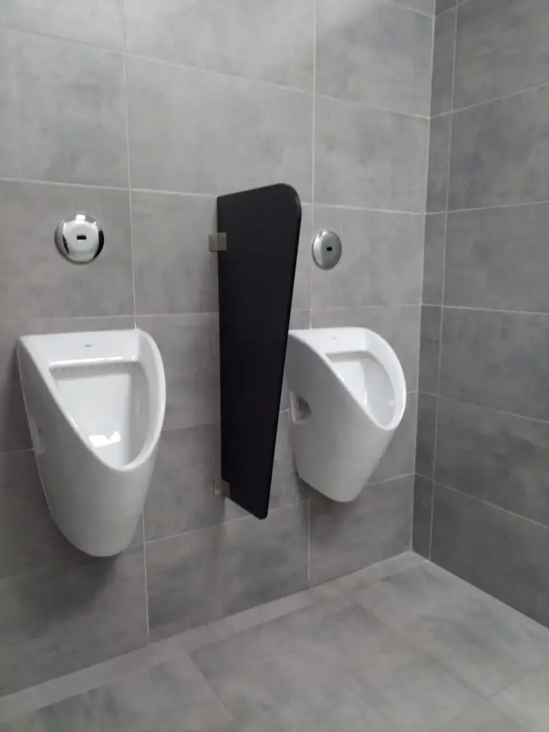 Separador de urinario de compacto fenólico negro, suspendido de la pared entre dos urinarios, en un aseo de azulejo gris