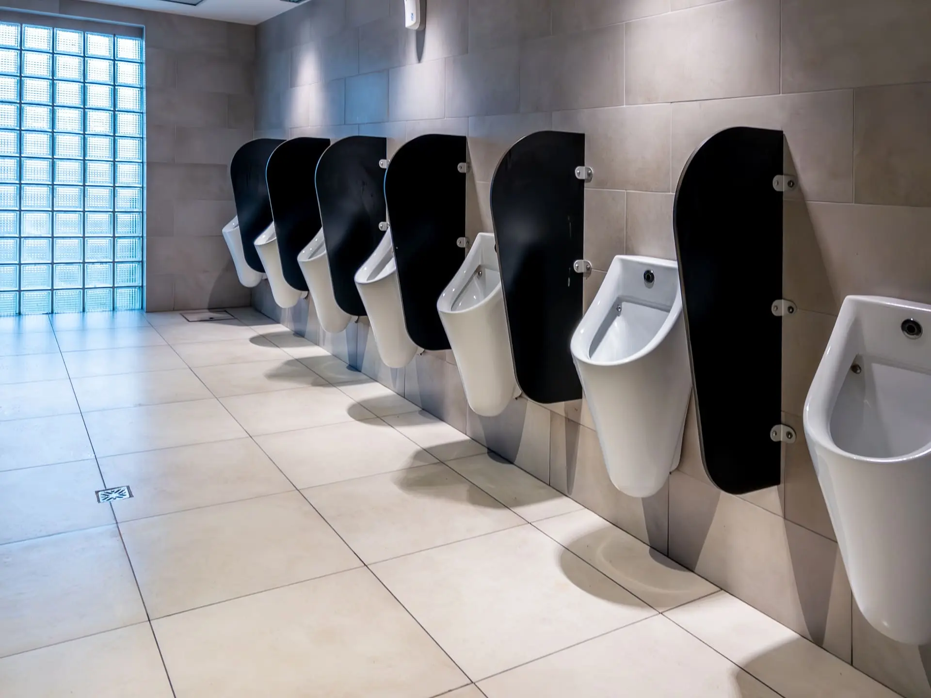 Urinarios con separadores fenólicos de color negro en un baño amplio y moderno, cada uno equipado con un sistema de descarga automática, bajo una iluminación natural filtrada - Bellum Logistics