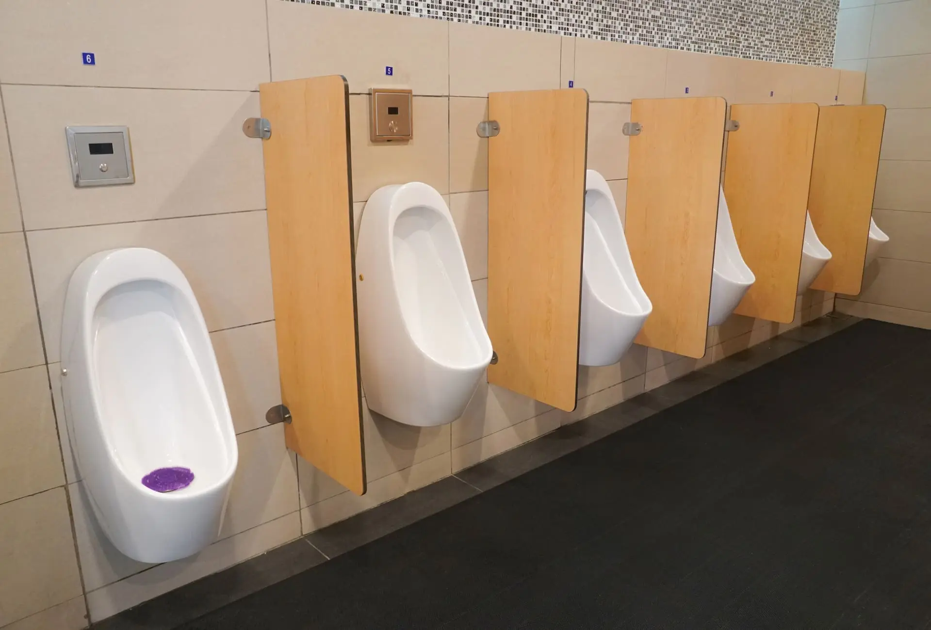 Fila de urinarios blancos separados por paneles fenólicos de madera clara montados sobre pinzas de metal, en un baño con azulejos y detalles modernos - Bellum Logistics