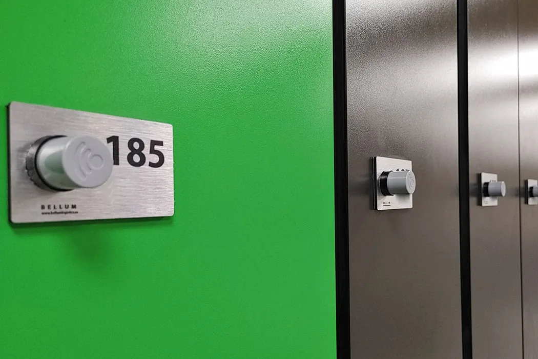 Placa numerada de acero cepillado con el número 185 alrededor de una cerradura RFID Ojmar OTS 10 Básica, en la puerta verde de una taquilla de gimnasio