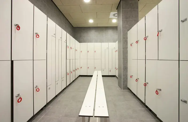 Vestuario de un gimnasio con taquillas fenólicas blancas a los dos lados y un banco en el centro