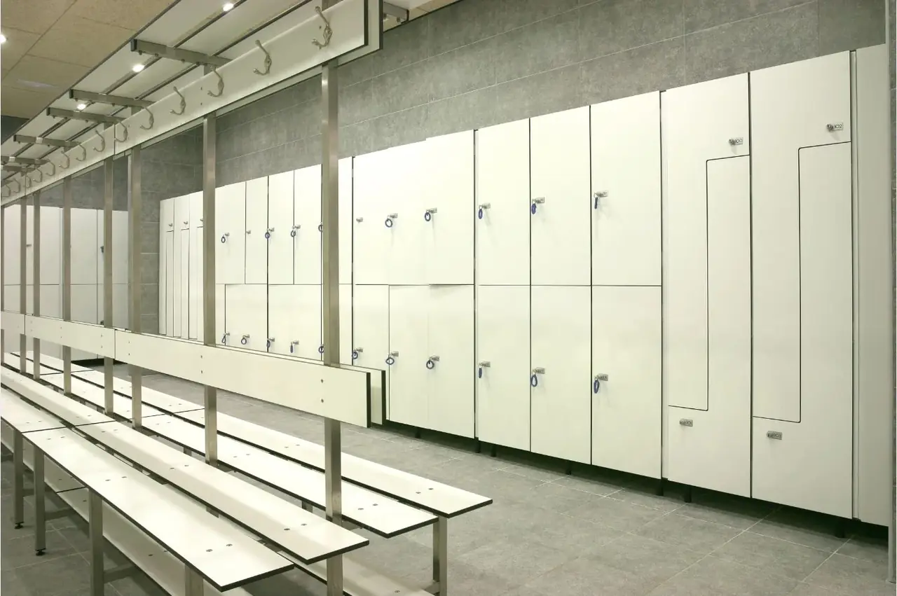 Taquillas fenólicas blancas de dos puertas y en L frente a bancos con colgadores