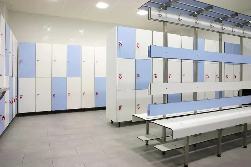 Vestuario con taquillas fenólicas de cuerpo blanco y puertas en azul y blanco, junto a un banco con colgadores