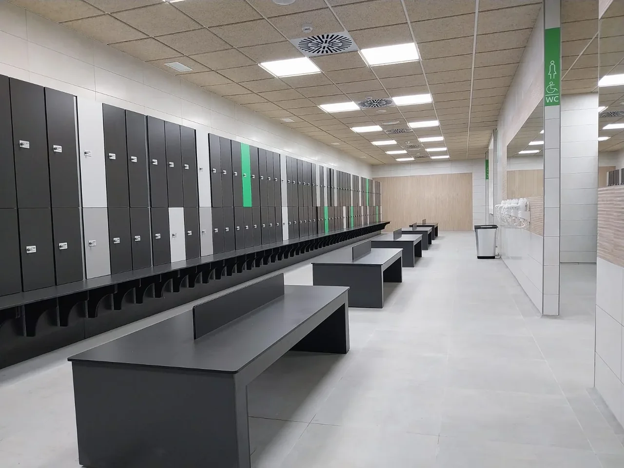 Vestuario de un gimnasio con una hilera larga de taquillas fenólicas en antracita, gris y verde con el banco incorporado, y bancos de compacto antracita en el centro